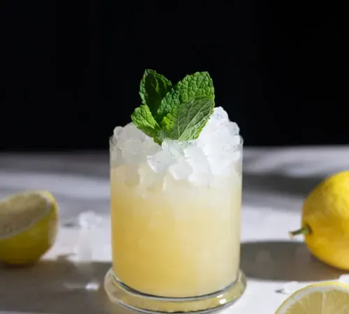 bebida amarilla sobre hielo picado en un vaso bajo con hojas de menta y limones.' title=