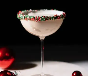 Bledý krémový nápoj v koktailovom pohári lemovanom červeným a zeleným posypom.' title=