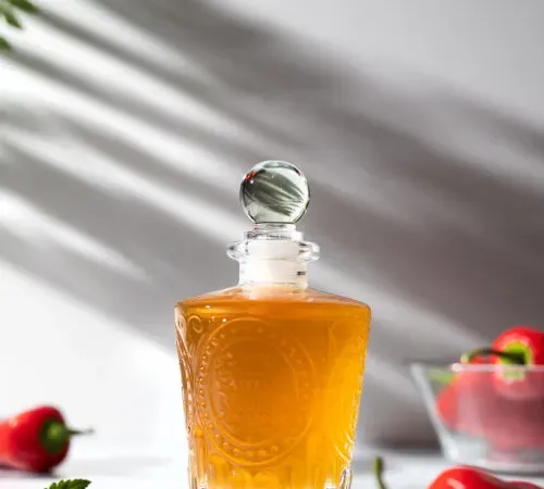 uma garrafa vintage cheia de líquido dourado rodeada de pimentão vermelho' title=