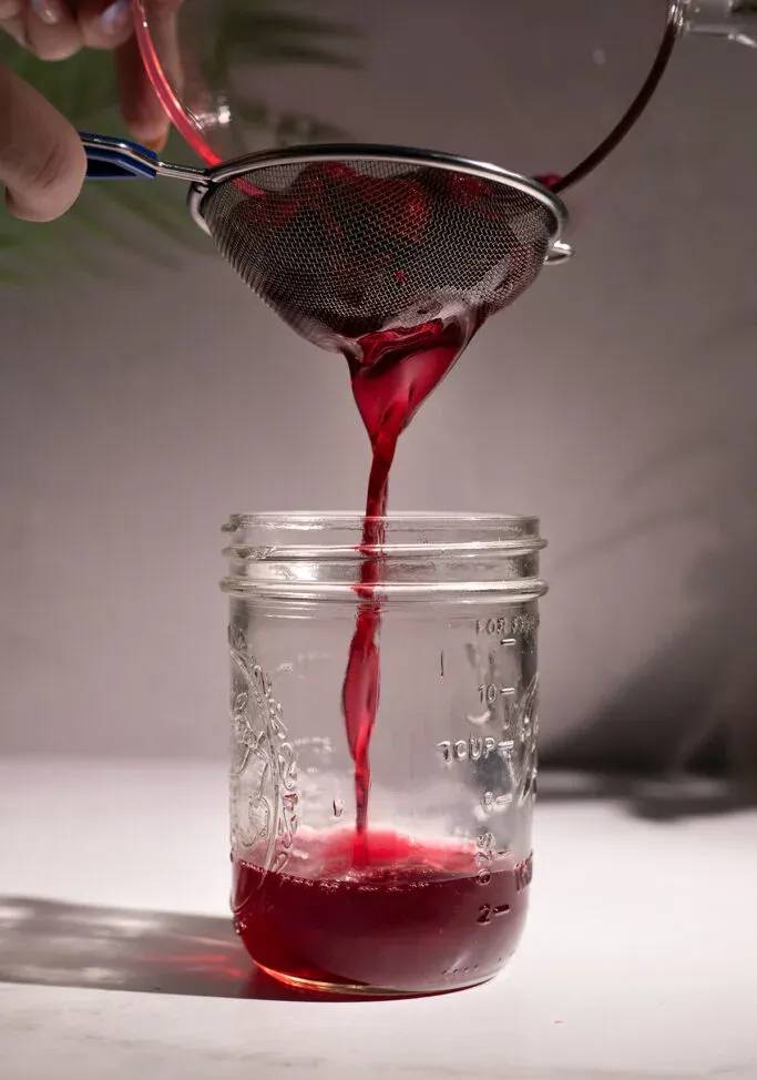 진한 빨간색 액체가 메쉬 스트레이너를 통해 유리병에 걸러지고 있습니다.' title=