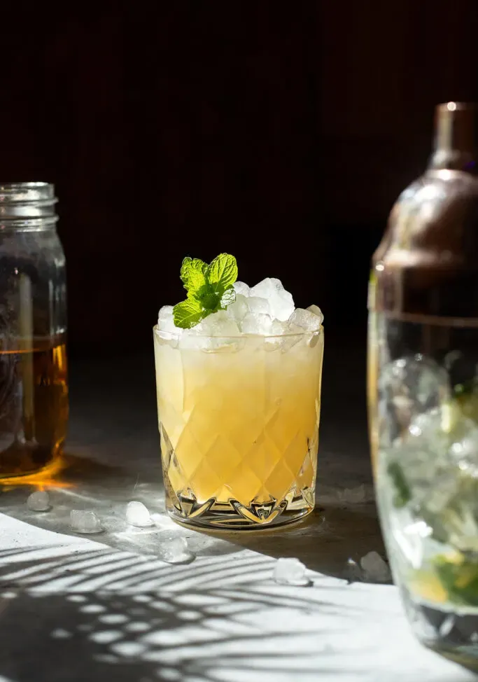 un cocktail dorato in un bicchiere rock accanto a uno shaker e un barattolo di liquido marrone.' loading='lazy' title=