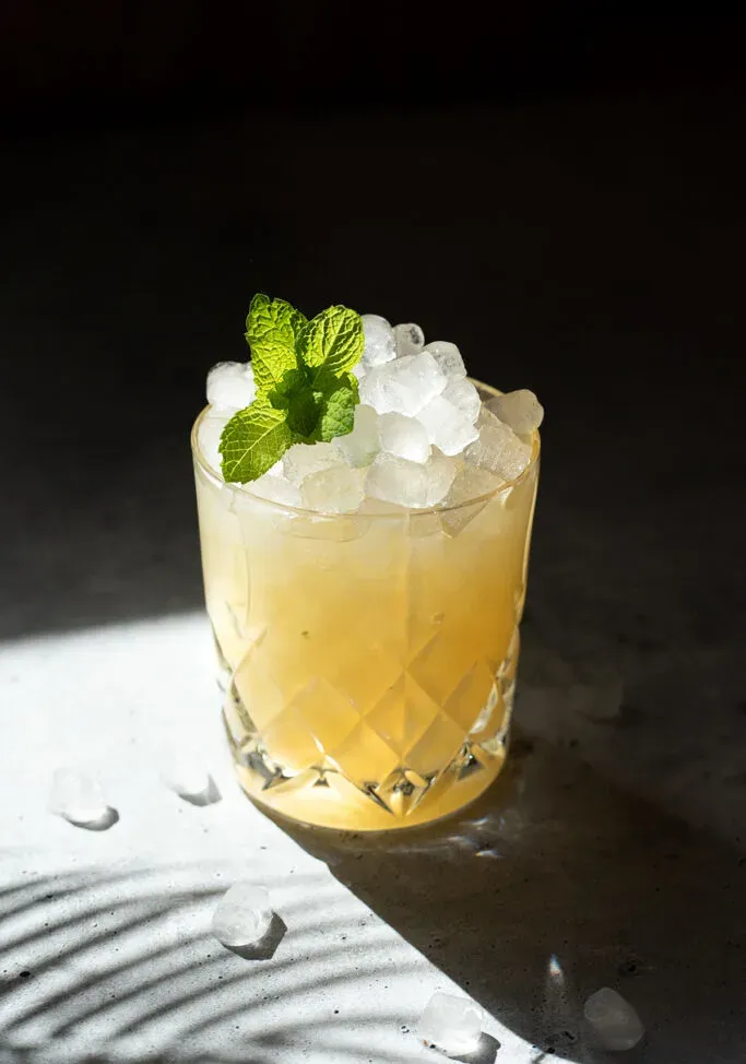 una bevanda gialla con ghiaccio tritato in un bicchiere rock con un rametto di menta.' title=