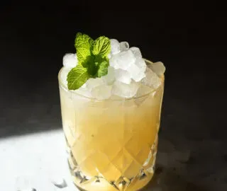 una bevanda gialla con ghiaccio tritato in un bicchiere rock con un rametto di menta.' title=