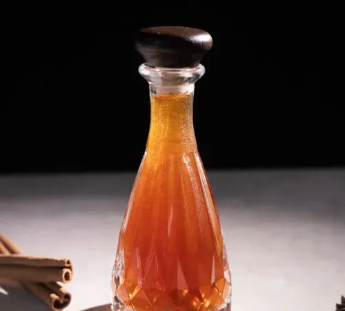 un'elegante bottiglia di vetro acidato piena di sciroppo di colore marrone aranciato.' title=