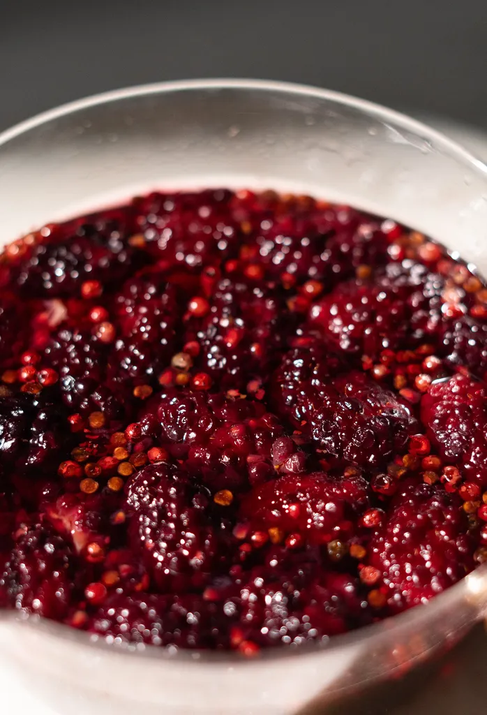 赤いシロップに入った調理されたブラックベリーと小さなピンクペッパーの接写。' loading='lazy' title=