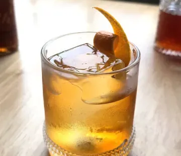 말린 오렌지 껍질 장식이 있는 로우볼 잔에 담긴 황금색 칵테일.' title=