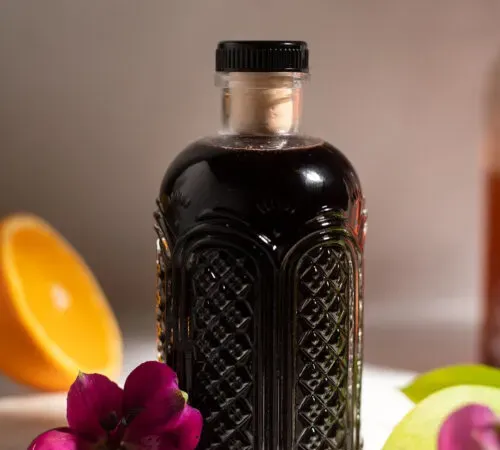 Dunkelvioletter Sirup in einer Glasflasche neben einer violetten Blume und einer Orangenscheibe.' title=