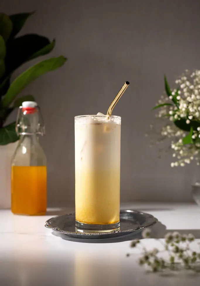 משקה צהוב קרמי גבוה עם קשית מתכת מוזהבת.' title=