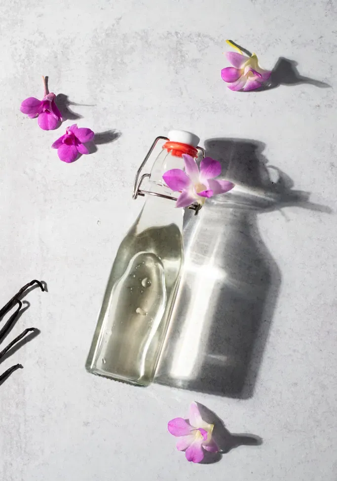 otočná fľaša číreho sirupu na bielom stole s ružovými orchideami.' loading='lazy' title=