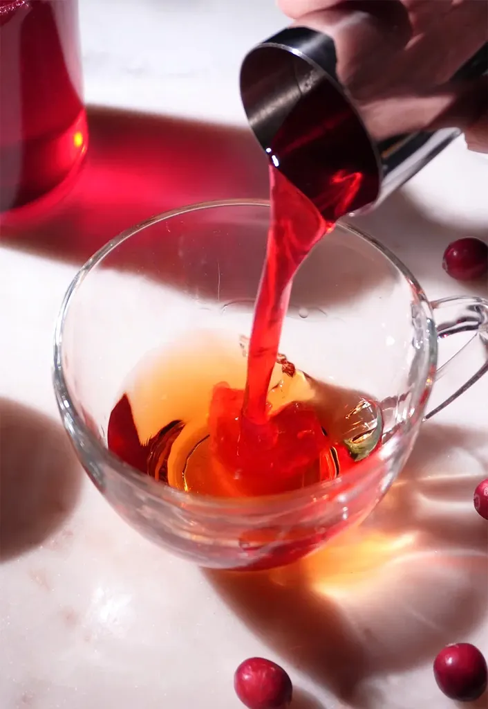 wlewanie syropu z czerwonej żurawiny z metalowego jiggera do szklanej filiżanki herbaty.' loading='lazy' title=