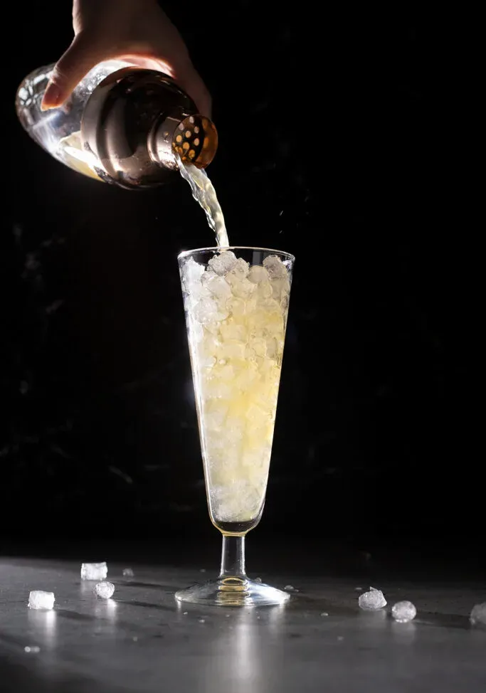 vertiendo una bebida de color amarillo pálido en un vaso alto lleno de hielo de guijarros.' loading='lazy' title=