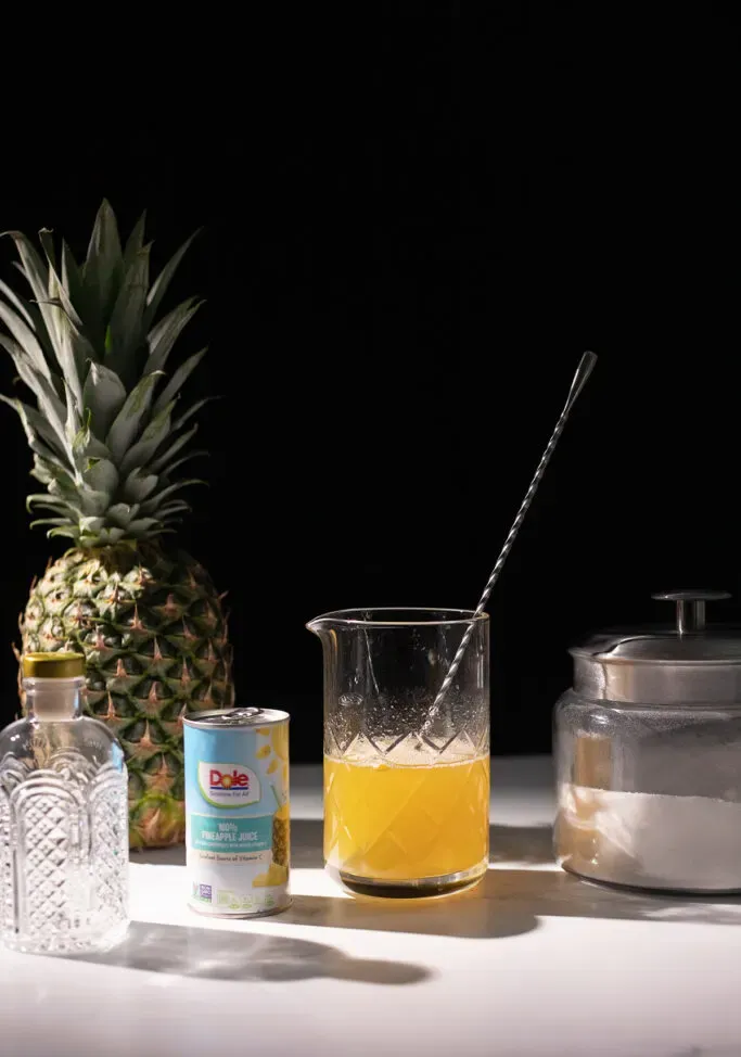 un ananas accanto a una brocca di sciroppo giallo e un contenitore di zucchero bianco.' title=