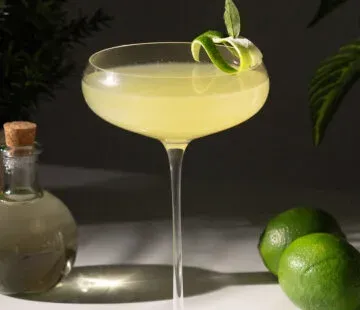 קוקטייל צהוב בכוס קופה גבוהה עם עיטור ליים ועלי דפנה על השפה.' title=