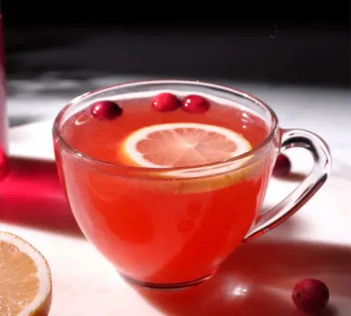 xícara de chá de vidro com líquido vermelho ao lado de cranberries e meio limão.' title=