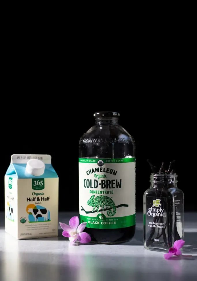 una bottiglia di concentrato di birra fredda accanto a un cartone di metà e metà e baccelli di vaniglia.' loading='lazy' title=