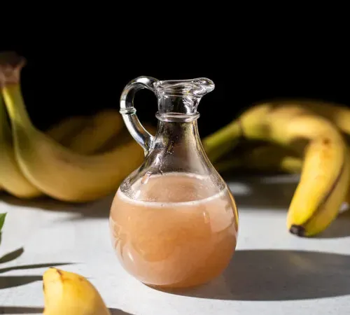 una jarra pequeña de almíbar espeso y dorado junto a plátanos.' title=