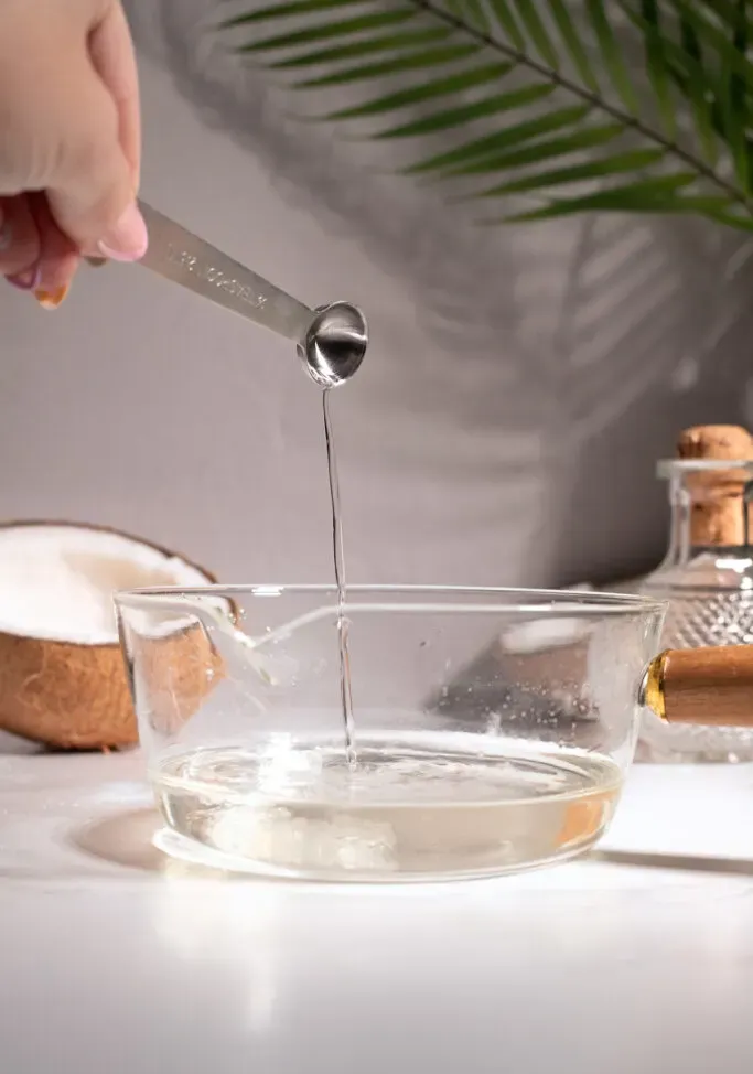 despejando extrato de coco transparente de uma colher de chá de metal em uma panela de vidro.' title=