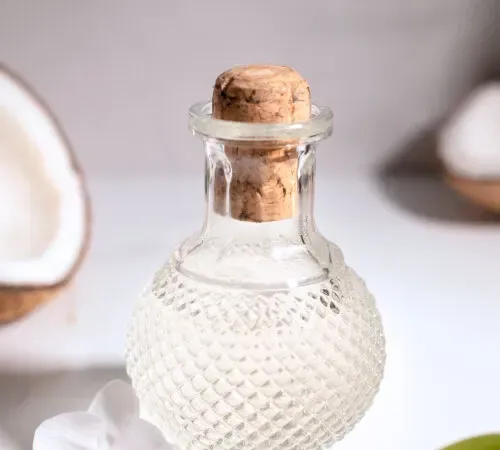 uma garrafa de vidro transparente vintage cheia de xarope de coco com uma orquídea e pedaços de coco.' title=