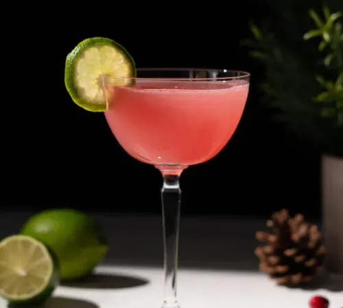 ライムホイールガーニッシュを添えたカクテルグラスに入った淡い赤色の飲み物。' title=