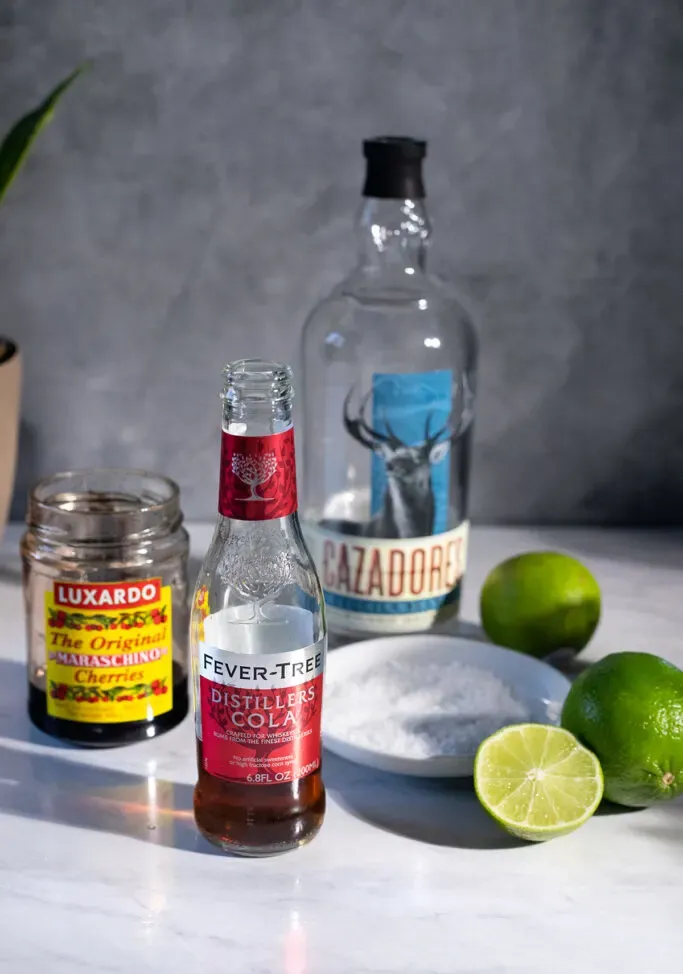 eine Flasche Tequila mit einem Glas Kirschen, eine Cola-Flasche, Limetten und Salz.' loading='lazy' title=