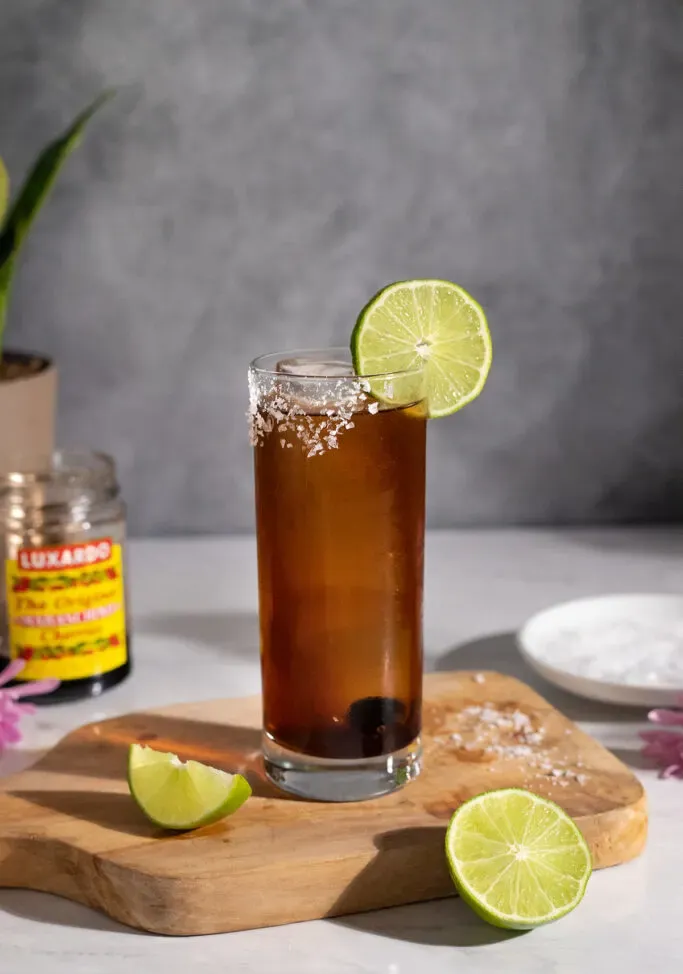 ein brauner Cocktail mit einem Limettenrad auf einem Holzschneidebrett.' loading='lazy' title=