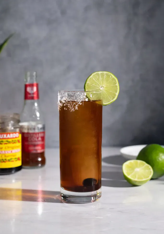 ein braunes Batanga-Getränk mit Cola-Flasche und Limetten.' title=