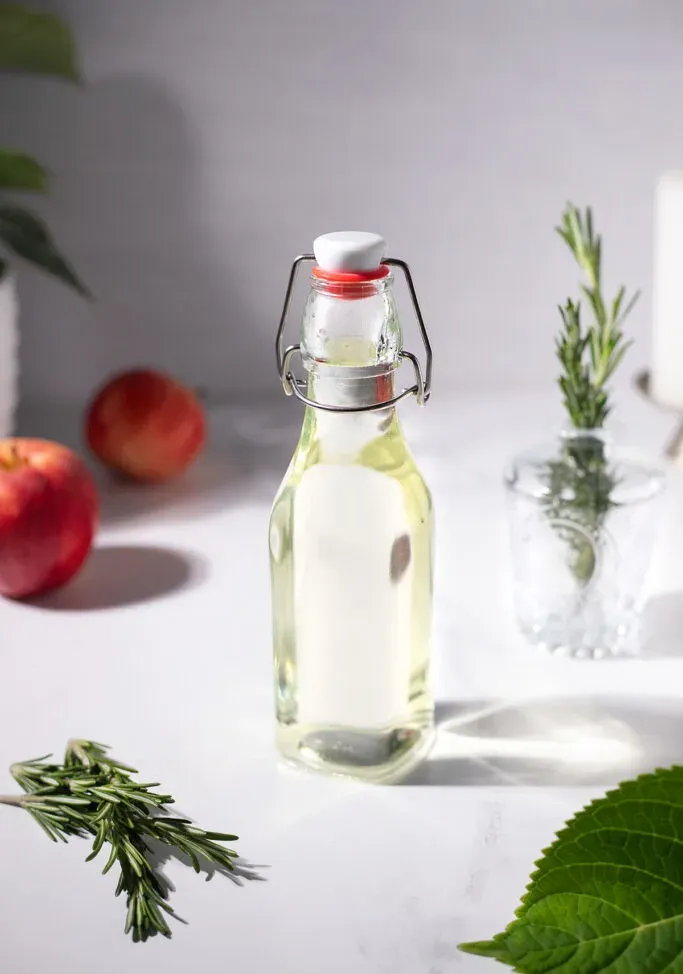 butelka z wahadłowym zamknięciem wypełniona jasnozielonym syropem obok gałązek rozmarynu i jabłek.' title=