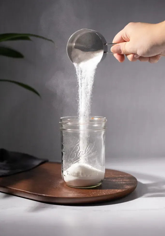 versare lo zucchero da un misurino in un barattolo di vetro.' loading='lazy' title=