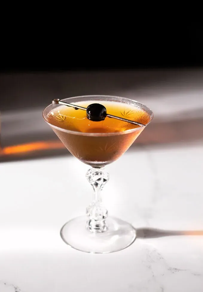 un cocktail marron dans un verre à cocktail vintage avec une cerise à cocktail.' title=