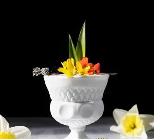 Tropischer Cocktail in einer weißen Glasschale mit Blumen und Ananasblättern.' title=