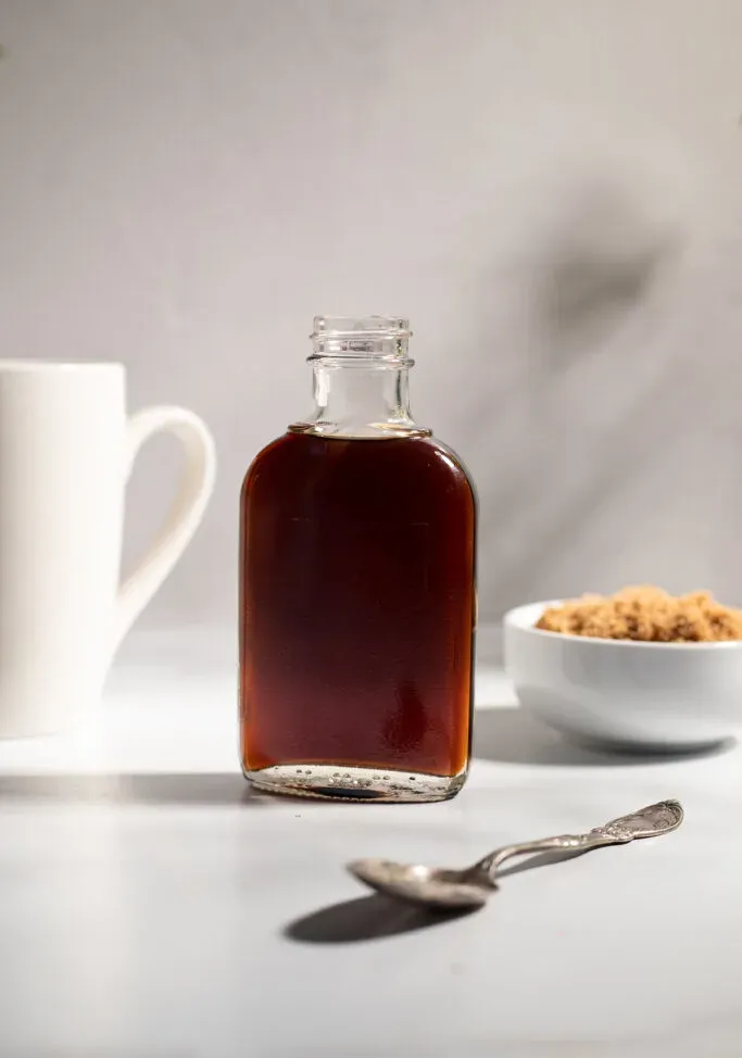 una piccola bottiglia di sciroppo marrone accanto a una tazza di caffè bianco.' title=