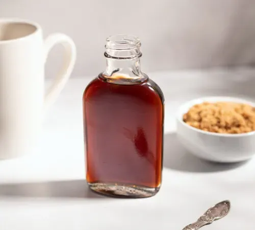 una piccola bottiglia di vetro di sciroppo marrone scuro accanto a una tazza di caffè bianco.' title=