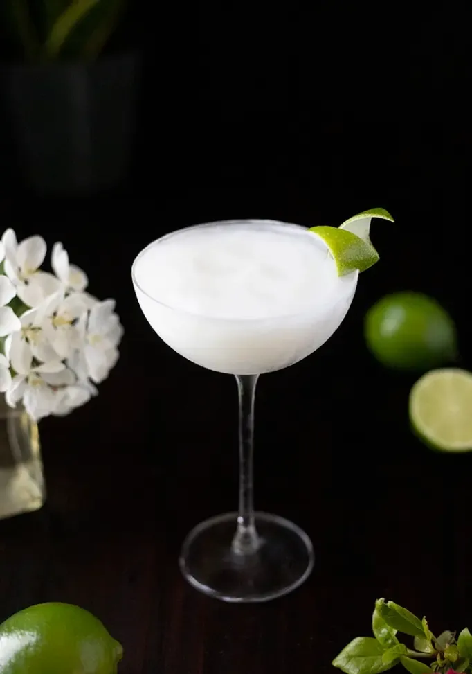 mrazený koktail v pohári kupé s dlhou stopkou na čiernom stole s limetkami a bielymi kvetmi' title=