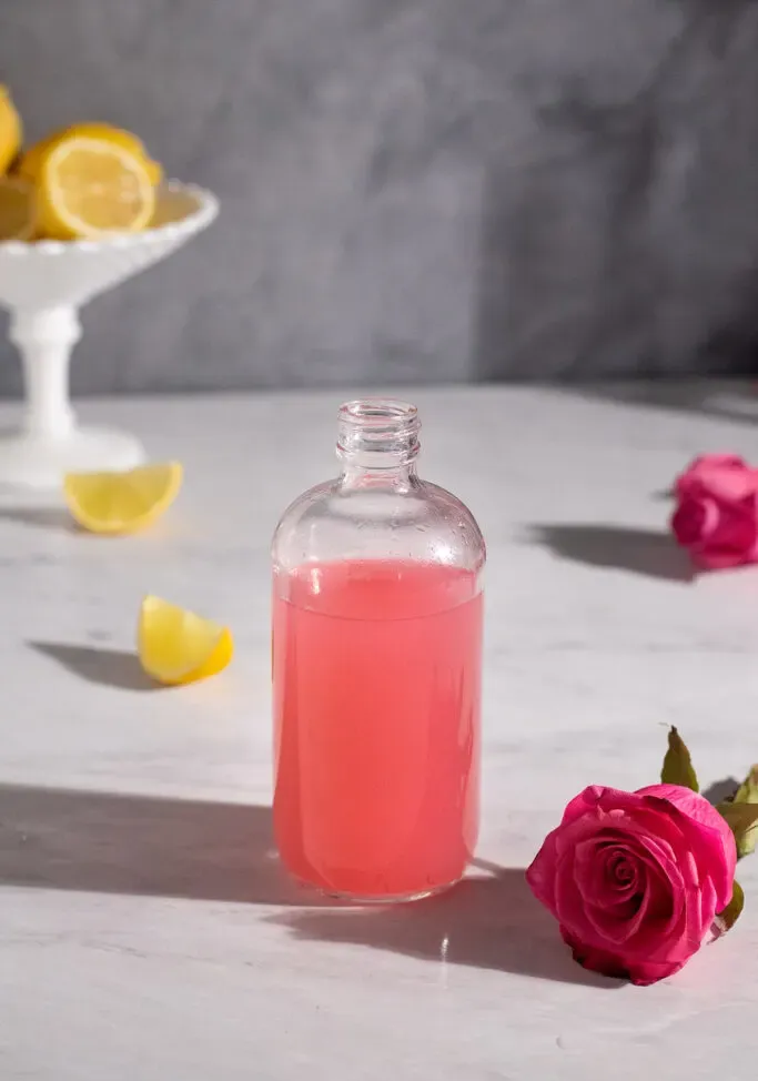 pequeña botella llena de jarabe rosa junto a una rosa rosa.' loading='lazy' title=