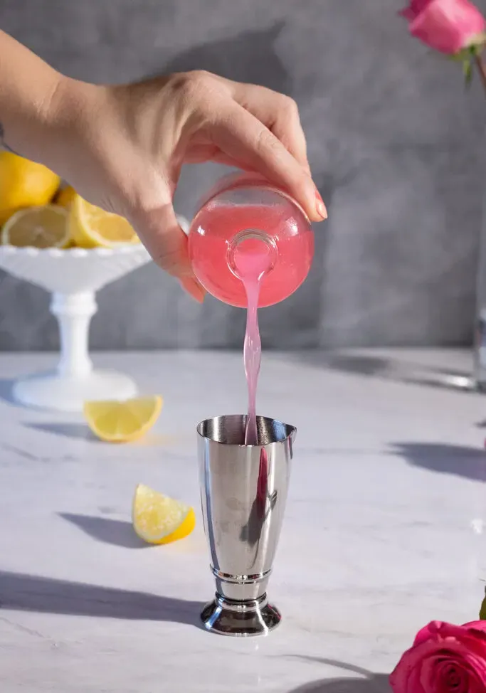 vertiendo almíbar rosado de una botella en una barra medidora de plata.' loading='lazy' title=