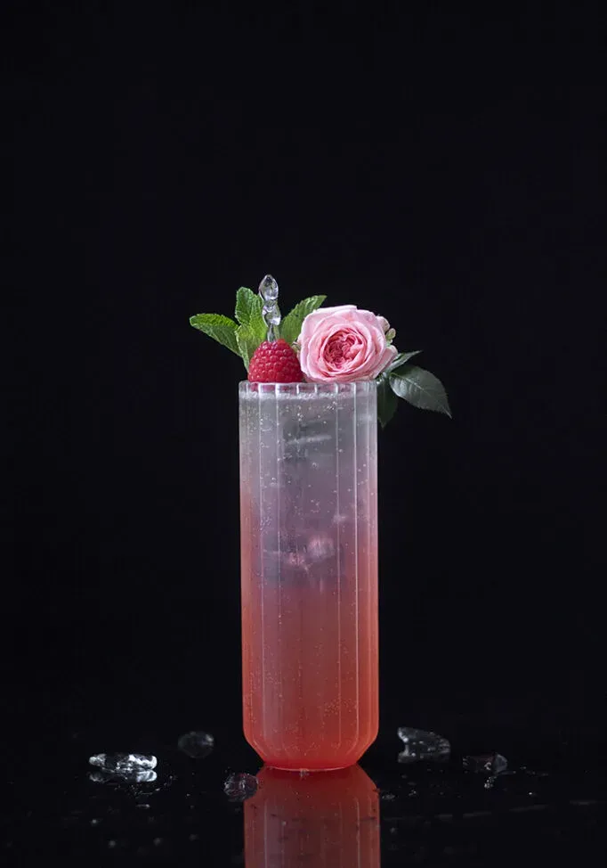 Vaso alto lleno de líquido rojo y adornado con frambuesa y rosa.' title=