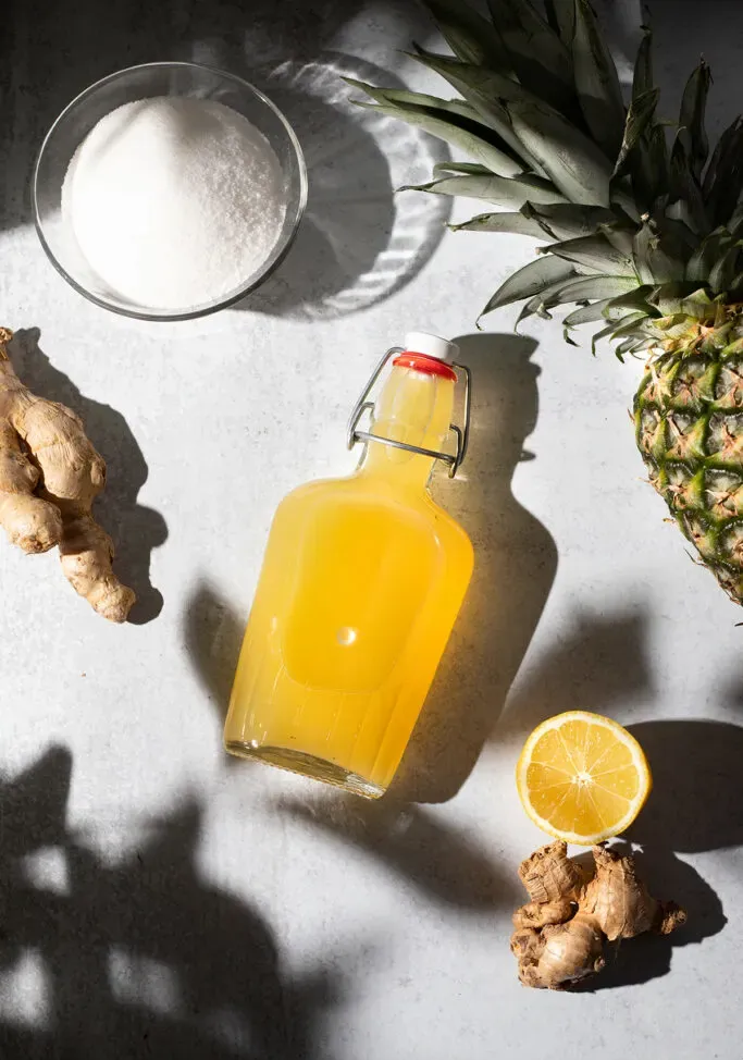 Flasche gelber Sirup auf einem weißen Tisch neben Zucker-Ingwer und einer Ananas.' loading='lazy' title=