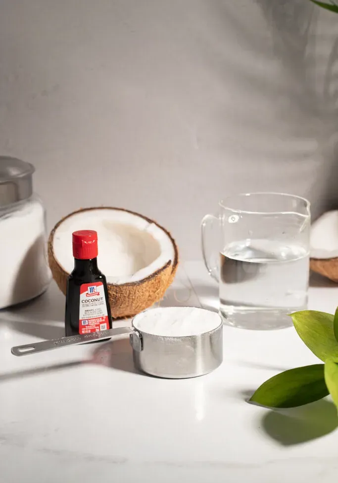 Kokosextrakt, eine Tasse Zucker, eine Tasse Wasser und eine halbe Kokosnuss auf einem weißen Tisch.' loading='lazy' title=