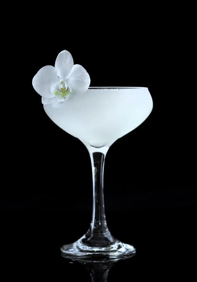 ein weißer Cocktail in einem Coupéglas, garniert mit einer weißen Orchidee.' title=