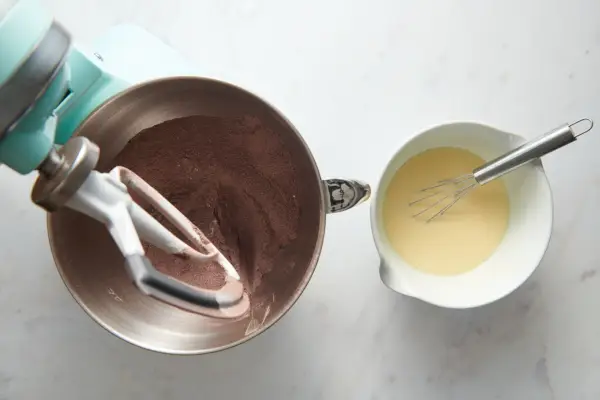 Un batteur sur socle avec de la farine, du sucre, de la poudre de cacao, du bicarbonate de soude, de la levure chimique et du sel à côté d'un petit bol de babeurre, d'huile, d'œufs et de vanille fouettés ensemble.