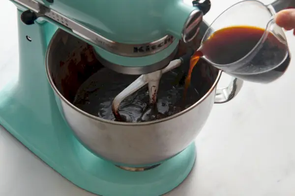 Une main versant une tasse à mesurer en verre de café chaud dans le bol d'un batteur sur socle pendant qu'il se mélange