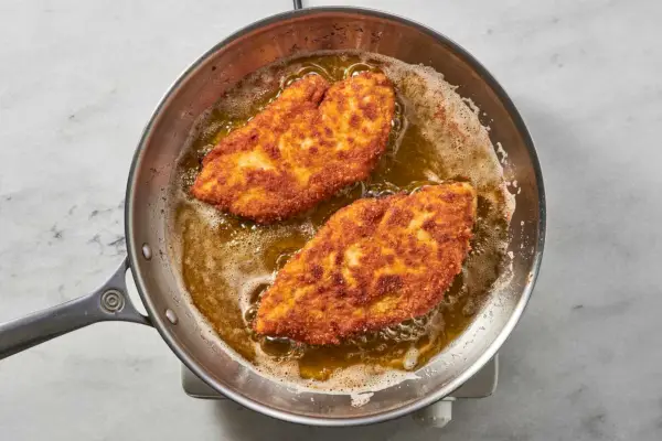 Eine Pfanne mit zwei goldbraun panierten Hähnchenbrustfilets, die in Öl gegart werden