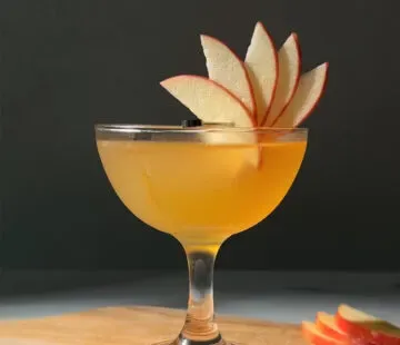 cocktail color oro in un bicchiere da coupé guarnito con un ventaglio di mele.' title=