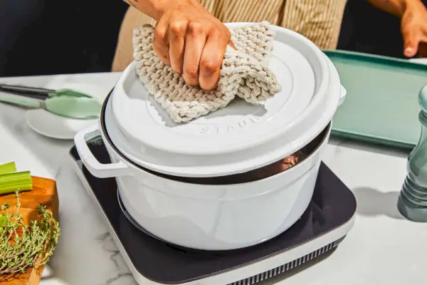 Person hebt den Deckel einer runden 5,5-Quart-Cocotte von Staub an und stellt sie mit den Zutaten auf den Tisch