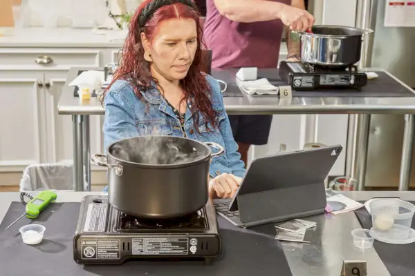 Kochendes Wasser in einem 10-teiligen Le Creuset Toughened Nonstick Pro Kochgeschirrset, während eine Person in der Nähe auf dem Laptop tippt