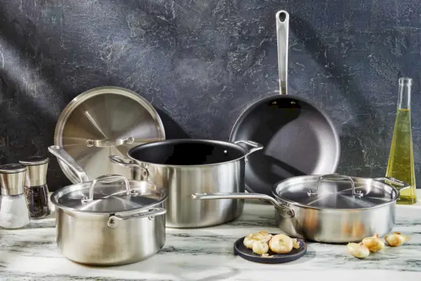 Hergestellt aus 7 Teilen. Das antihaftbeschichtete Set wird auf einer Marmorarbeitsplatte mit Salz- und Pfefferstreuern, Knoblauch, Zwiebeln und Öl angezeigt