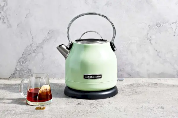 Czajnik elektryczny KitchenAid ustawiony na marmurowym blacie obok szklanego kubka z torebką herbaty w środku