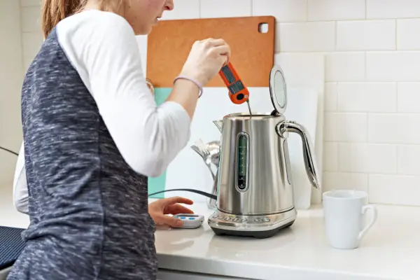 Osoba wkładająca termometr do czajnika Breville Smart Kettle Luxe, wystawiona na białym blacie z pobliskim białym kubkiem
