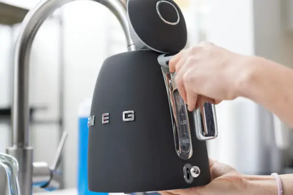 Dłoń trzymająca uchwyt czajnika elektrycznego Smeg podczas napełniania wodą z kranu kuchennego