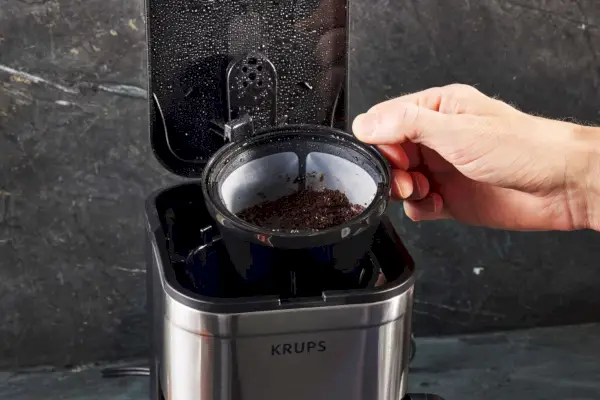 Close-up de uma mão retirando borra de café usada da cafeteira Krups Simply Brew de aço inoxidável, 5 xícaras